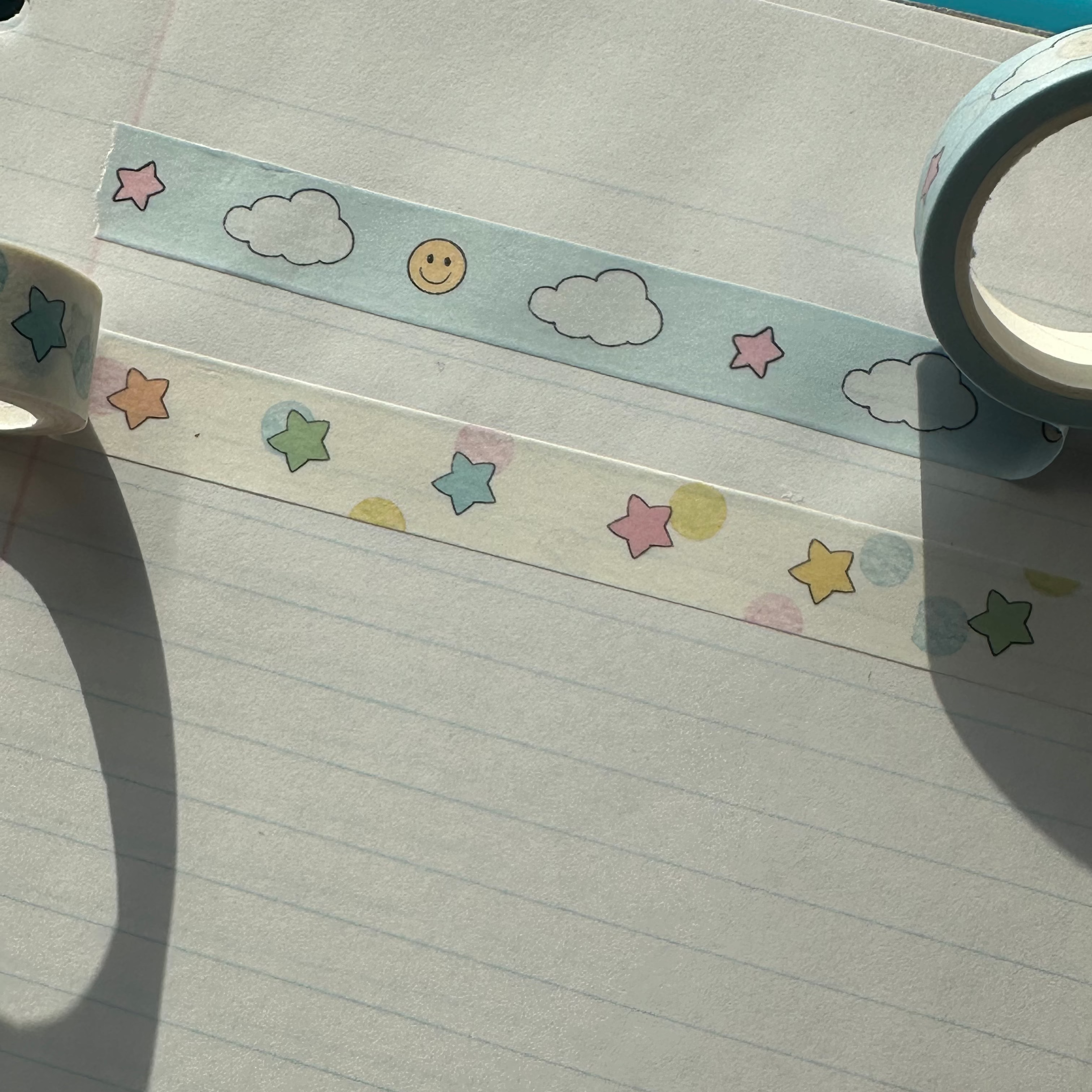 Two rolls of decorative washi tape with pastel star and cloud patterns laid out on lined notebook paper under direct sunlight.